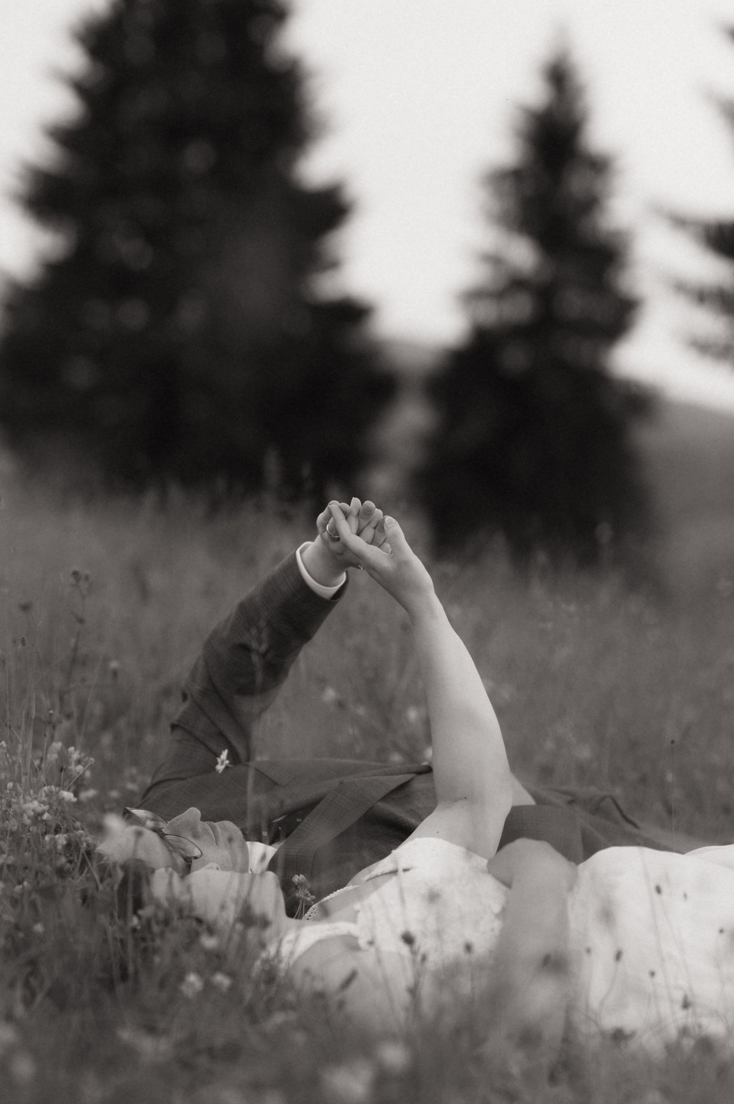 Black and white photo of couple lying in meadow grass holding hands up, with blurred evergreen trees in background.