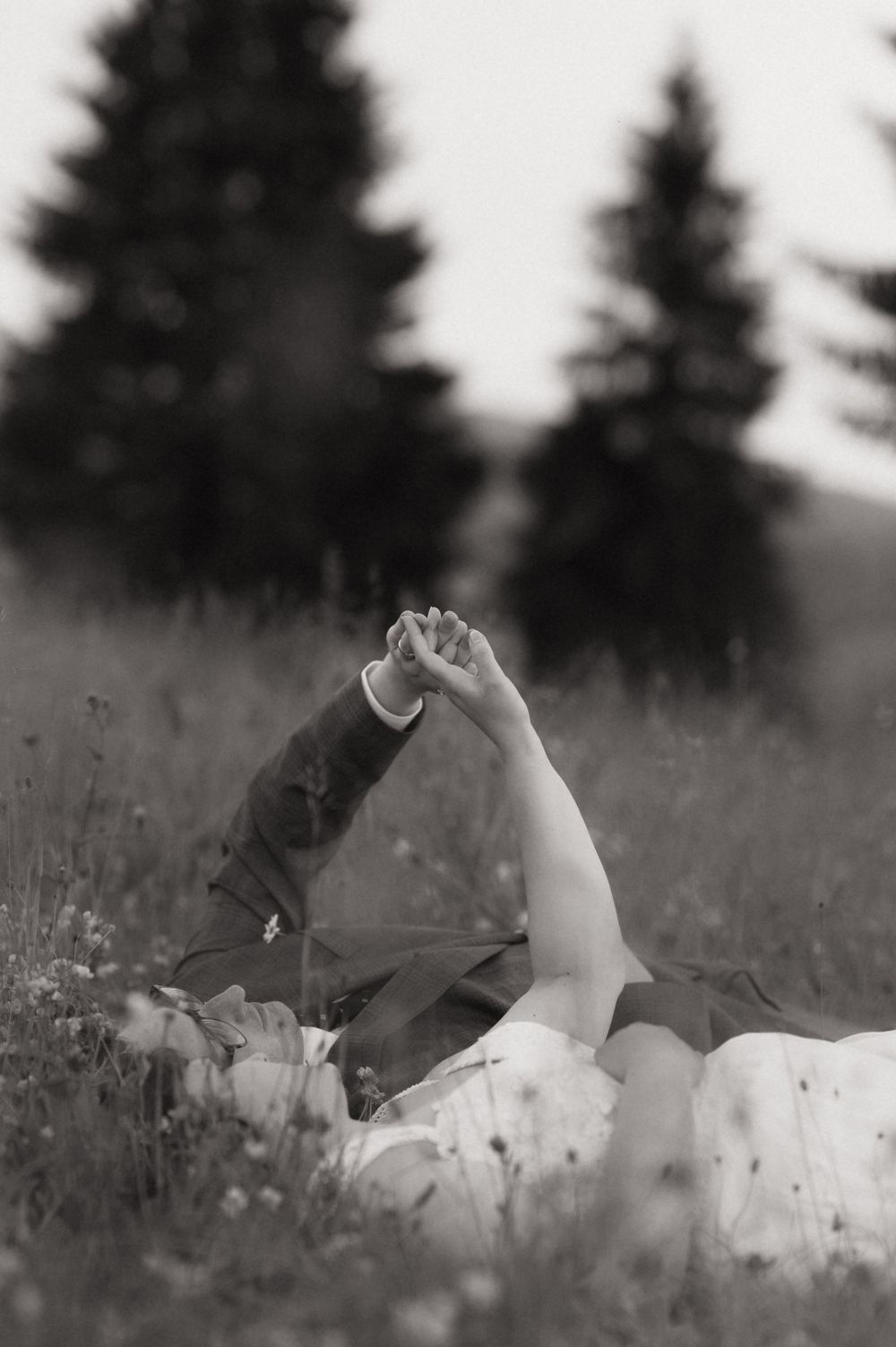 Black and white photo of couple lying in meadow grass holding hands up, with blurred evergreen trees in background.