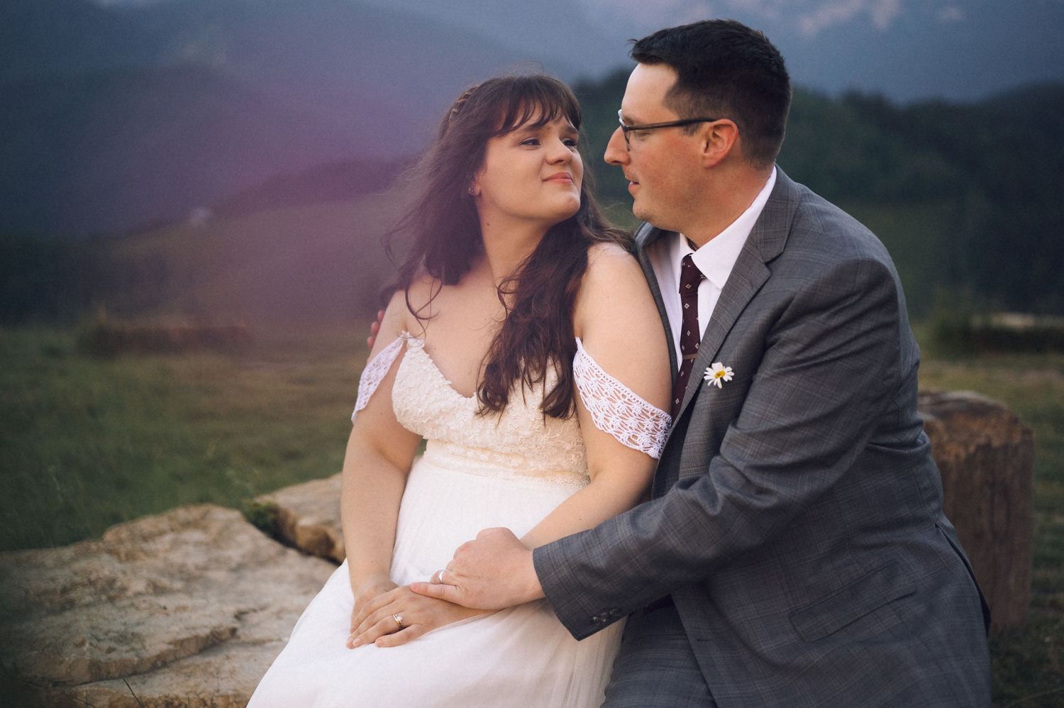 Couple in wedding attire sitting on rocks with mountain backdrop, groom in gray suit, bride in white dress.