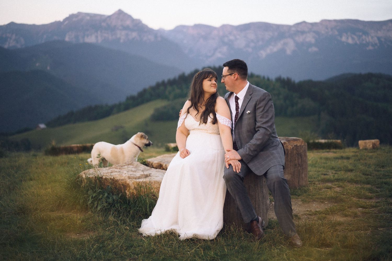 Couple shares romantic moment on hay bale with pug dog, mountain landscape backdrop at outdoor wedding.