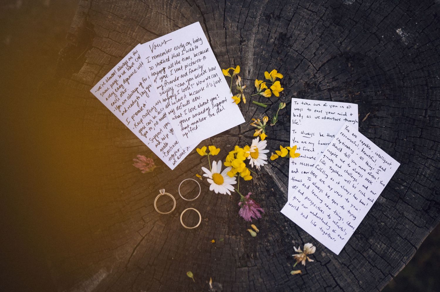 Handwritten vow cards with wildflowers and gold wedding rings on rustic wood stump, flat lay wedding detail photo.