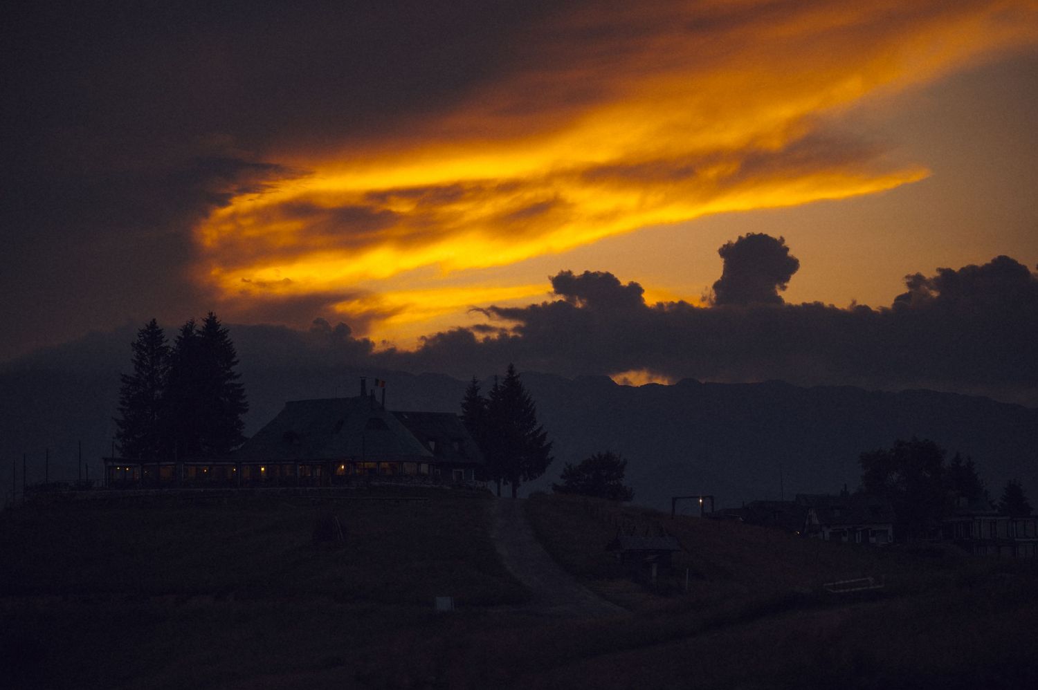 Dramatic sunset with fiery orange clouds silhouetting a rural farmhouse and trees against a dark stormy sky.
