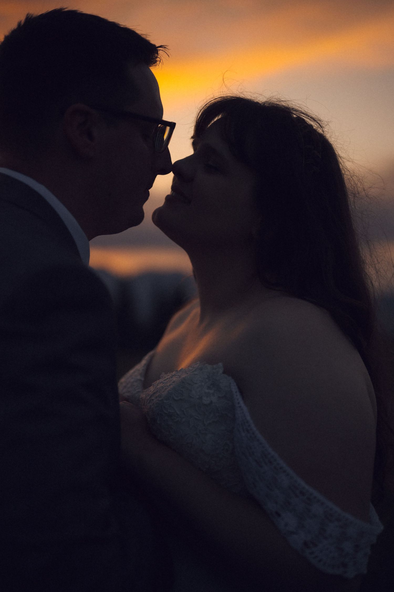 Romantic silhouette of a bride and groom sharing a kiss at sunset with golden orange sky backdrop.