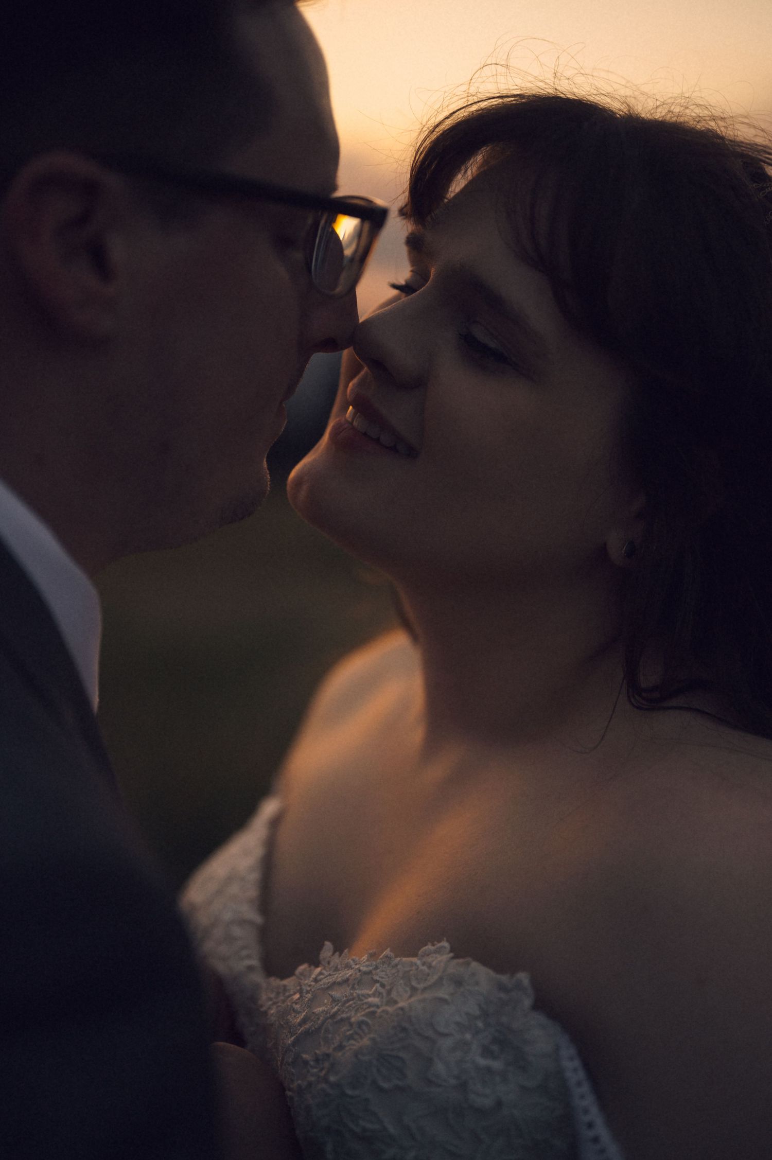 A bride and groom share a romantic almost-kiss at sunset, with warm golden light illuminating their faces.