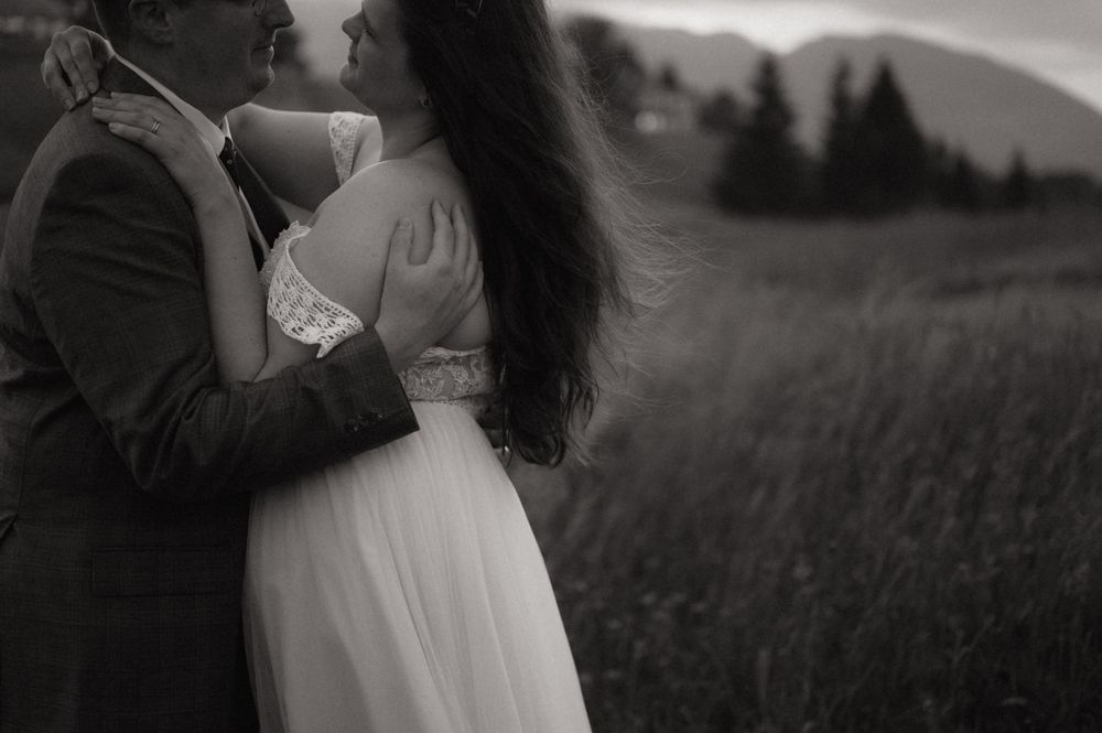 Black and white photo of a couple embracing in a field, the bride in a white gown with mountains in the background.