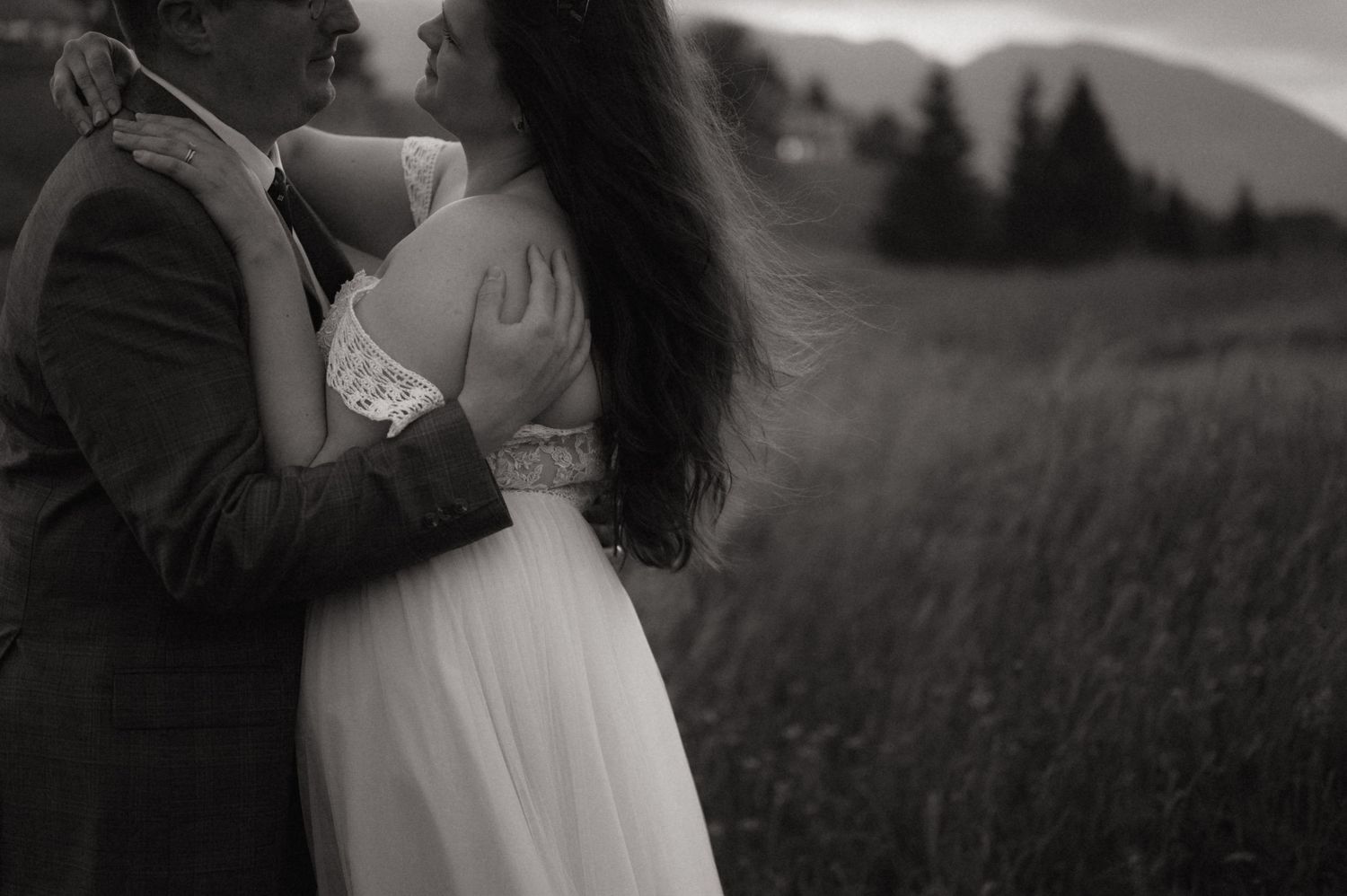 Black and white photo of a couple embracing in a field, the bride in a white gown with mountains in the background.