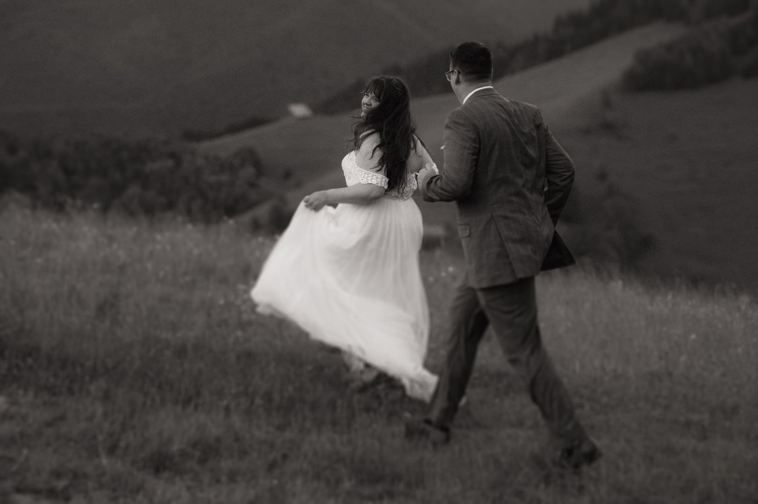 Black and white photo of a bride and groom walking through a lush green hillside meadow on their wedding day.