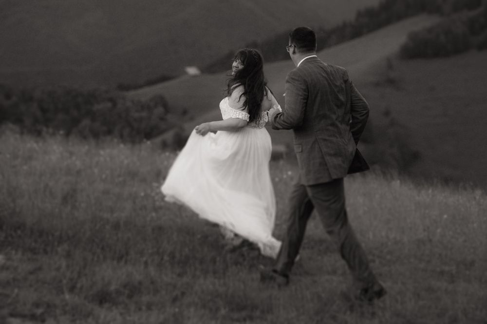 Black and white photo of a bride and groom walking through a lush green hillside meadow on their wedding day.