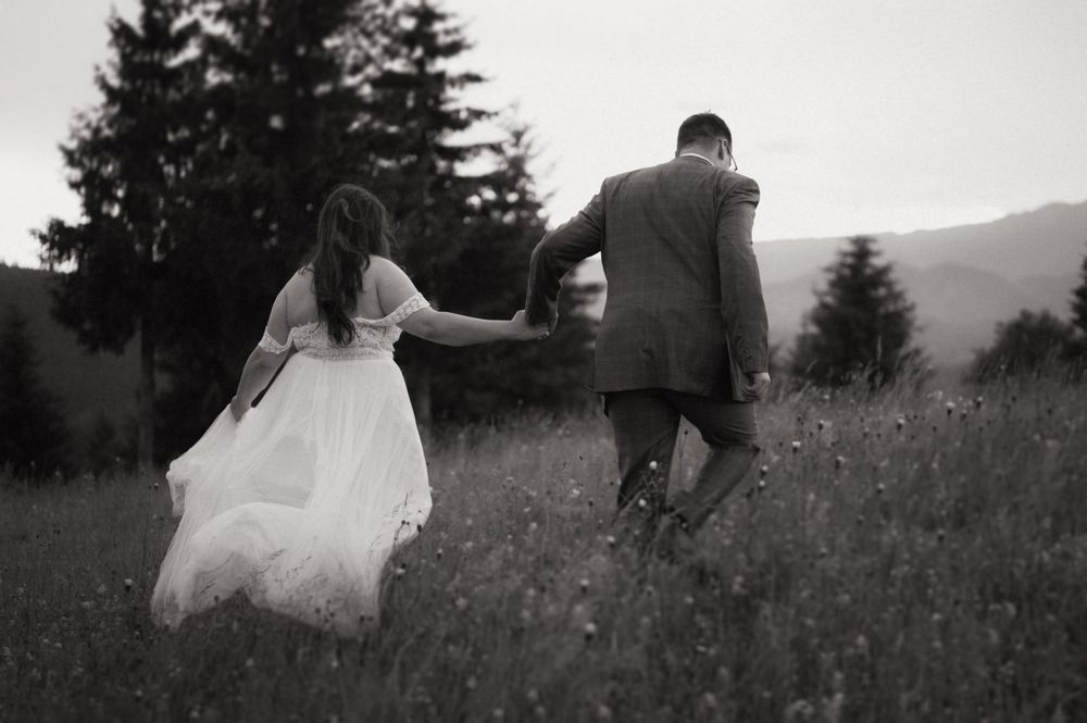 Black and white photo of a bride and groom holding hands walking through a wildflower meadow with mountains in the background.