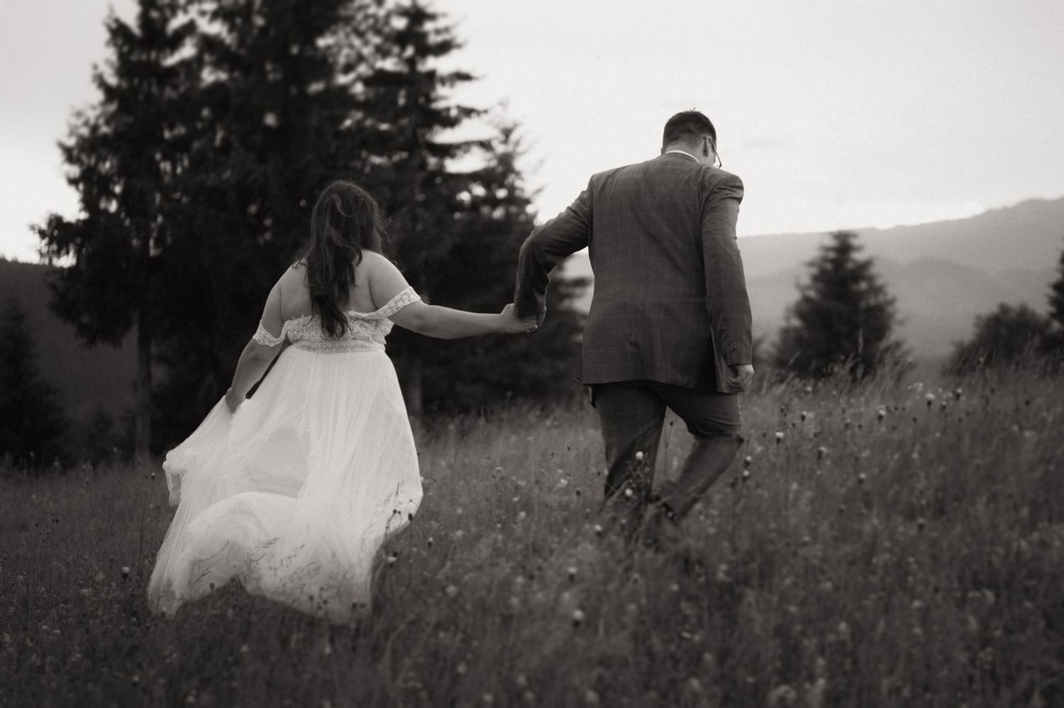 Black and white photo of a bride and groom holding hands walking through a wildflower meadow with mountains in the background.