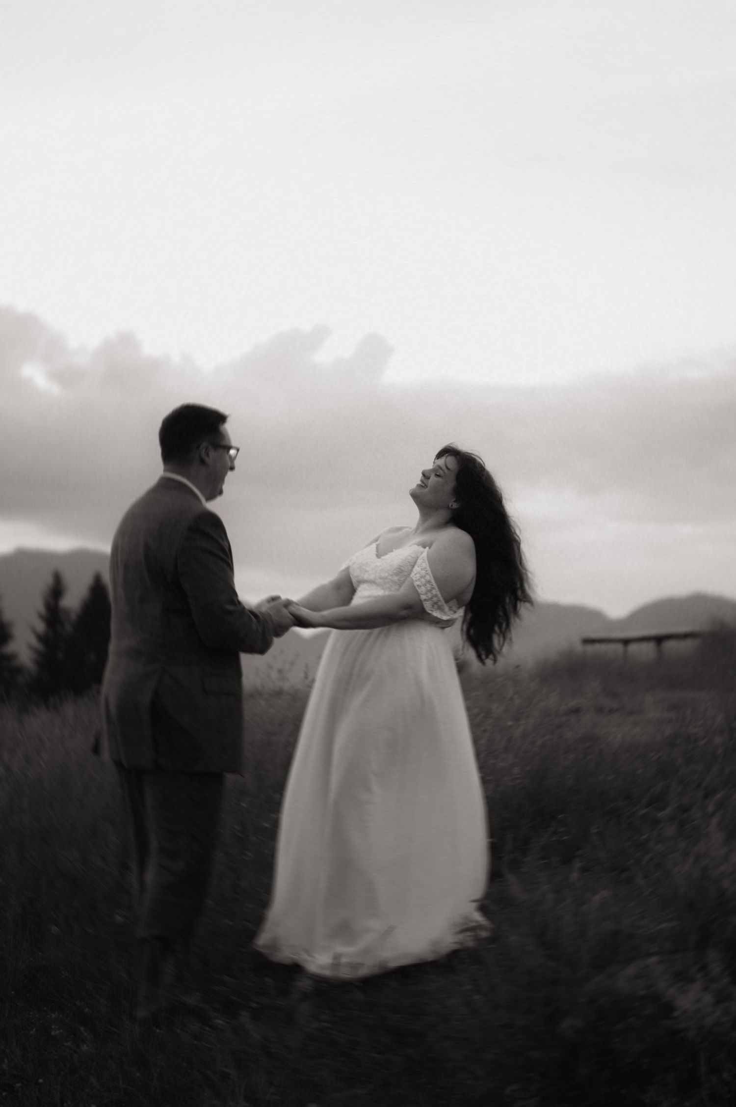 Black and white photo of a couple holding hands in a field, bride in white dress laughing joyfully under cloudy skies.