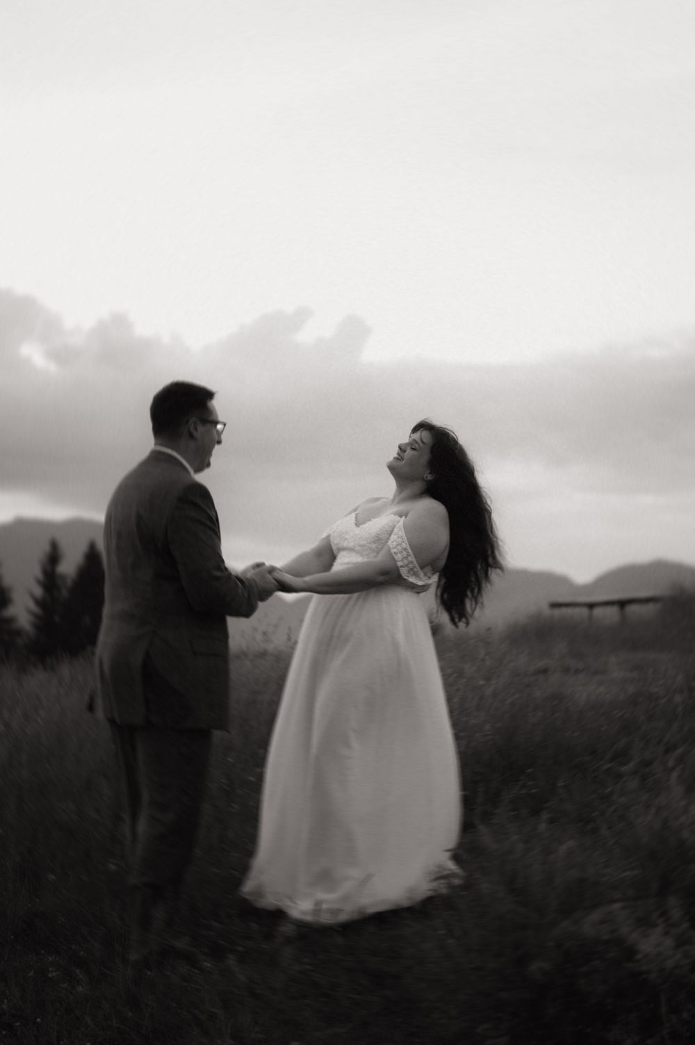 Black and white photo of a couple holding hands in a field, bride in white dress laughing joyfully under cloudy skies.