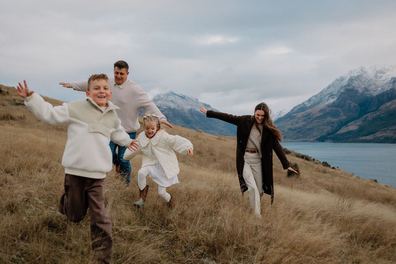 Family of four playing in golden grass fields with snow-capped mountains and lake backdrop in New Zealand.