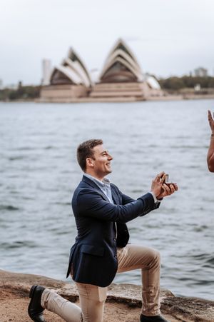 Man proposing on one knee with engagement ring in front of iconic Sydney Opera House waterfront.