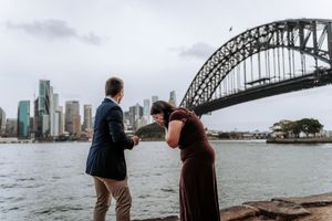 Couple sharing a proposal moment at Sydney Harbour Bridge waterfront, man presenting ring to emotional woman.