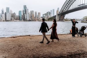 Couple walking hand in hand along Sydney Harbour foreshore with Harbour Bridge and city skyline backdrop.