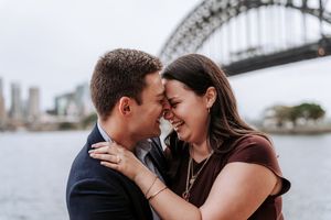 Couple sharing a romantic moment with Sydney Harbour Bridge in the background on a cloudy day.