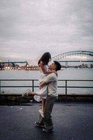A couple shares a romantic kiss as the man lifts the woman near a waterfront with a bridge at dusk.