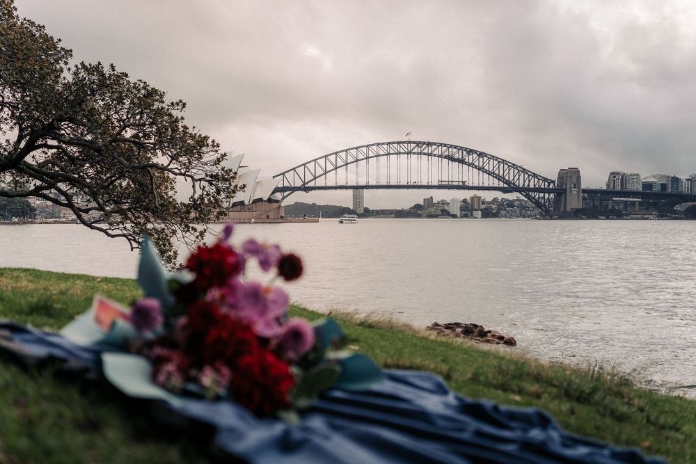 Bouquet of red and purple flowers on a blanket by Sydney Harbour Bridge on a cloudy day.