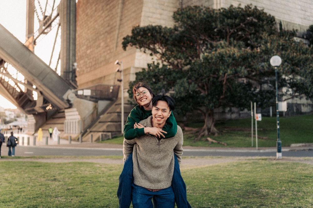 A joyful couple shares a piggyback ride on green grass near the Sydney Harbour Bridge on a sunny day.