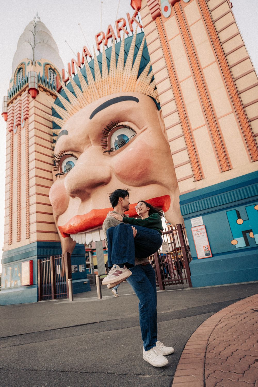 Couple embracing at Luna Park Sydney entrance, featuring the iconic giant face, in a romantic outdoor photo.