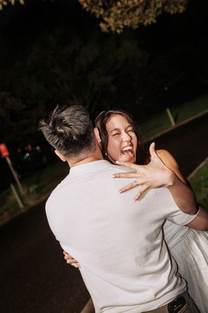 Joyful woman laughing and hugging a man outdoors at night, showing off an engagement ring on her hand.