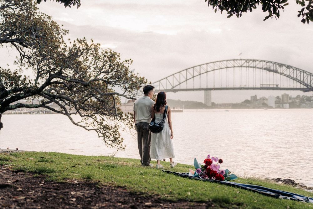 Couple standing together on grassy waterfront admiring Sydney Harbour Bridge view with colorful flowers nearby.