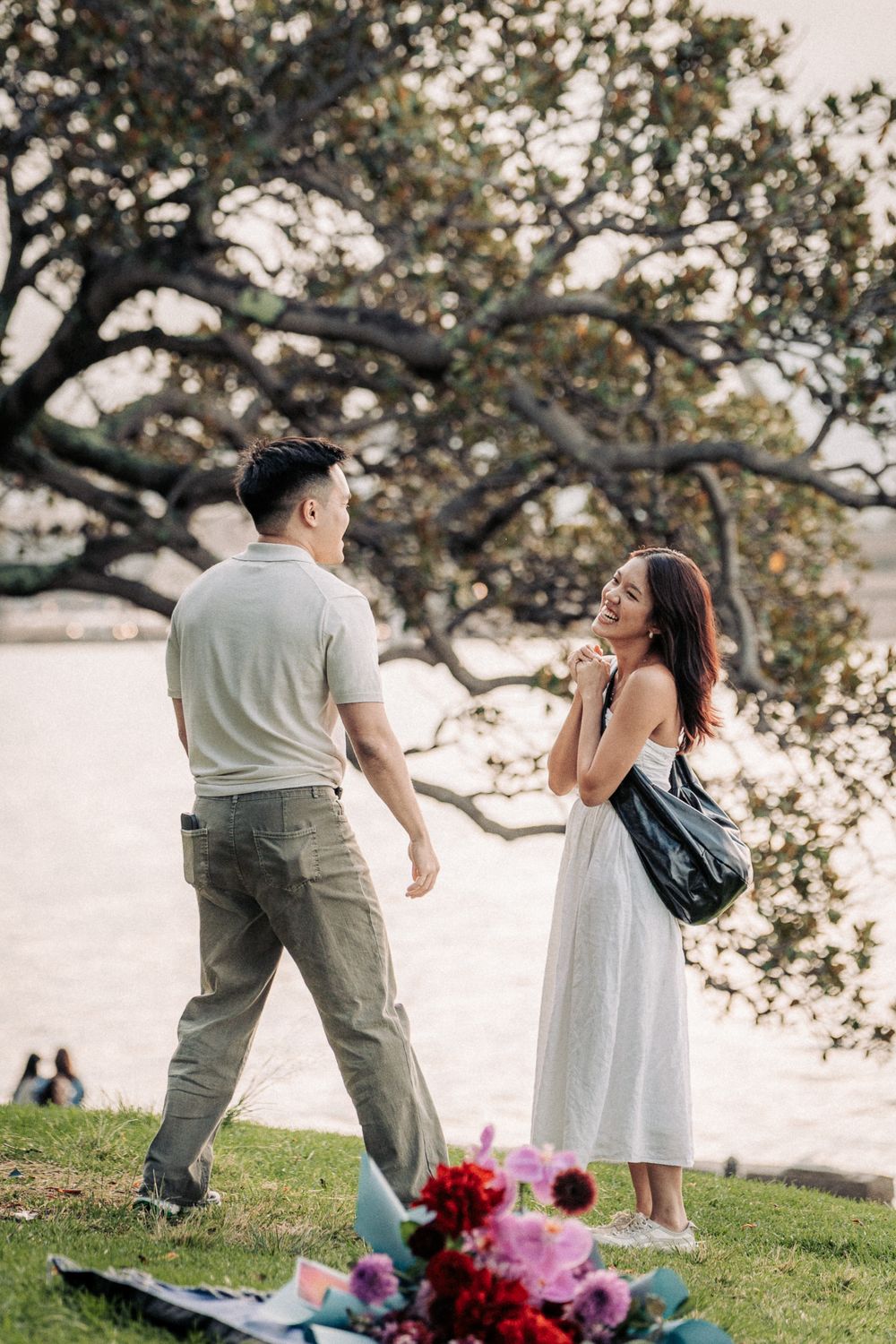 Couple sharing a romantic moment by the waterfront with flowers in the foreground and a large tree backdrop.