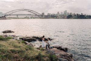 Couple sitting on rocky shore with Sydney Harbour Bridge and city skyline in the background.