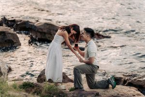 Man proposes to woman on rocky shoreline at sunset, kneeling with ring box as waves crash behind them.