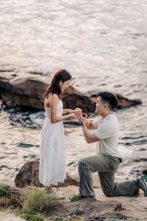Man proposes to woman on rocky shoreline, kneeling with ring box as she smiles in white dress.