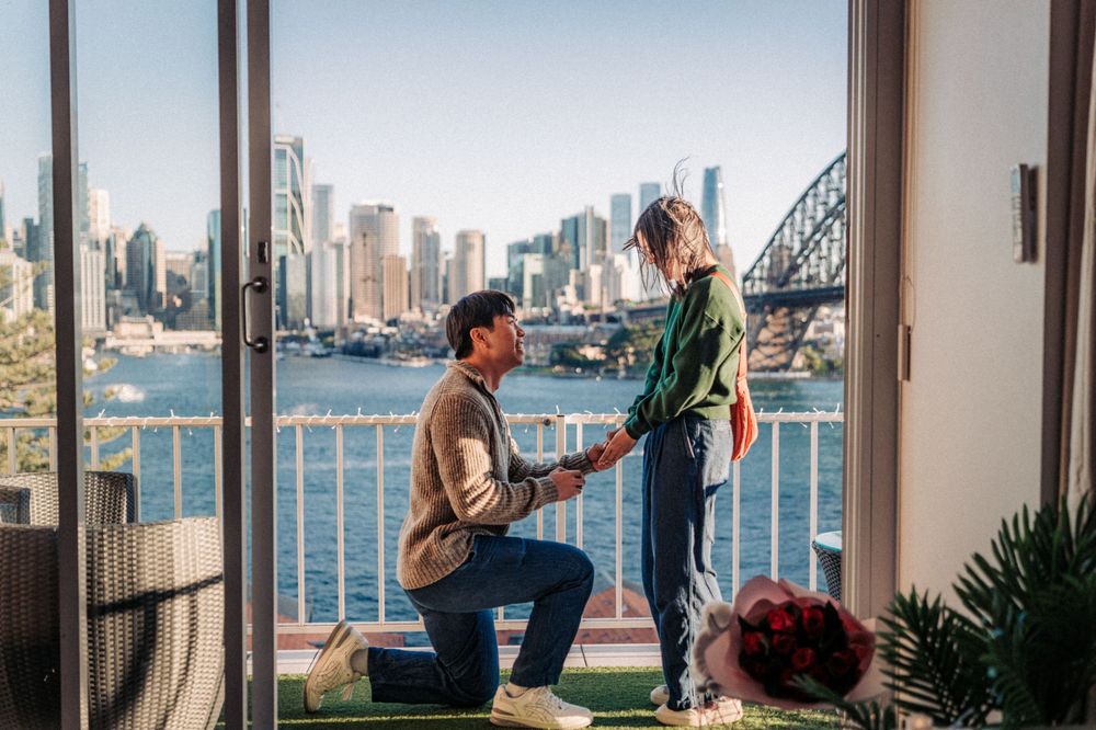 Man proposing to woman on balcony with Sydney Harbour Bridge and city skyline in background.