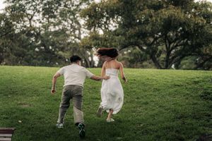 Couple in wedding attire running hand-in-hand across a lush green park, bride in white gown, groom in casual outfit.