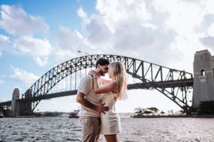 Couple sharing a romantic kiss in front of the iconic Sydney Harbour Bridge on a sunny day.