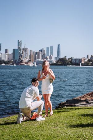 Man proposes to woman on grass by Sydney Harbour with Opera House and city skyline in background.