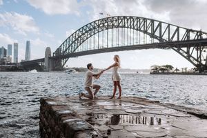 Man proposes to woman on rocky waterfront with Sydney Harbour Bridge backdrop on a cloudy day.