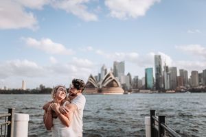 Couple embracing on Sydney Harbour waterfront with Opera House and city skyline in background on a sunny day.