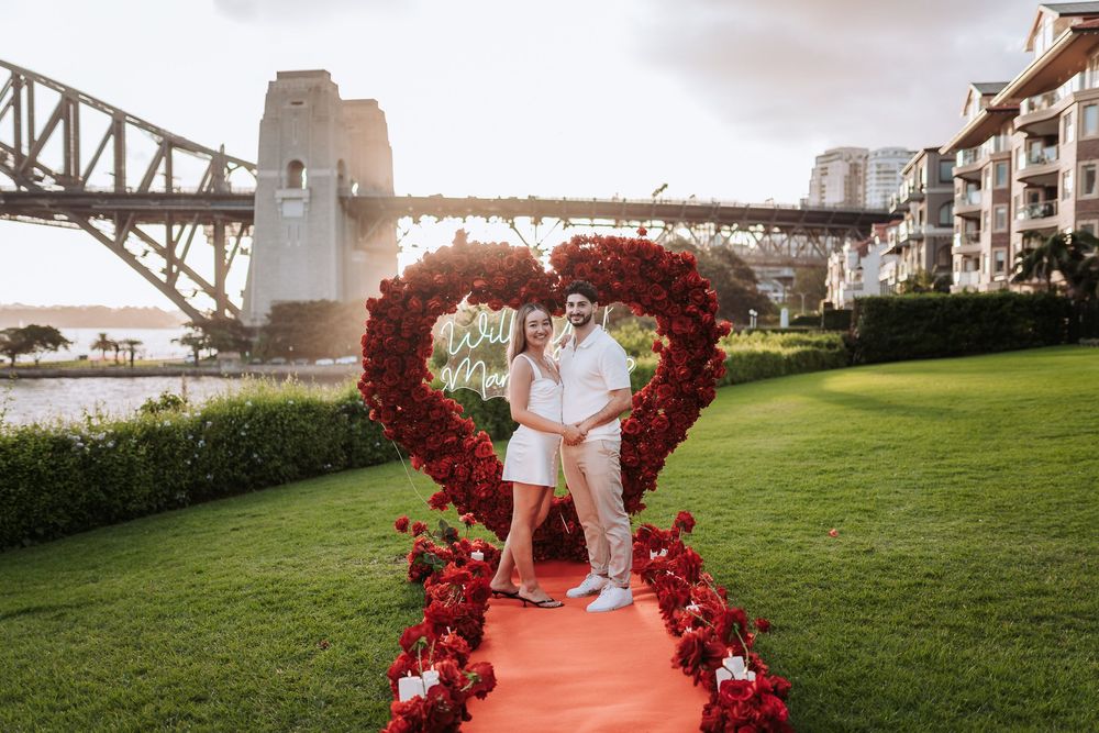 Couple poses under red rose heart arch on green lawn with Sydney Harbour Bridge backdrop during romantic proposal setup.