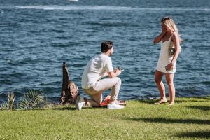 Man kneeling on grass proposing to woman in white dress by the water on a sunny day.