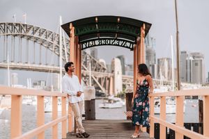 Couple standing on a wooden pier with Sydney Harbour Bridge in the background, man in white and woman in floral dress.