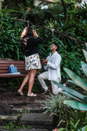Man proposes on one knee to woman in lush garden setting, holding ring box as she reacts with surprise.