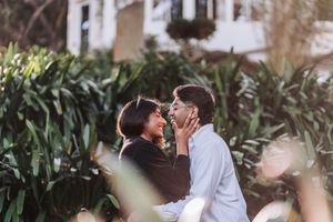 Romantic couple sharing an intimate moment surrounded by lush green tropical plants and a white house backdrop.