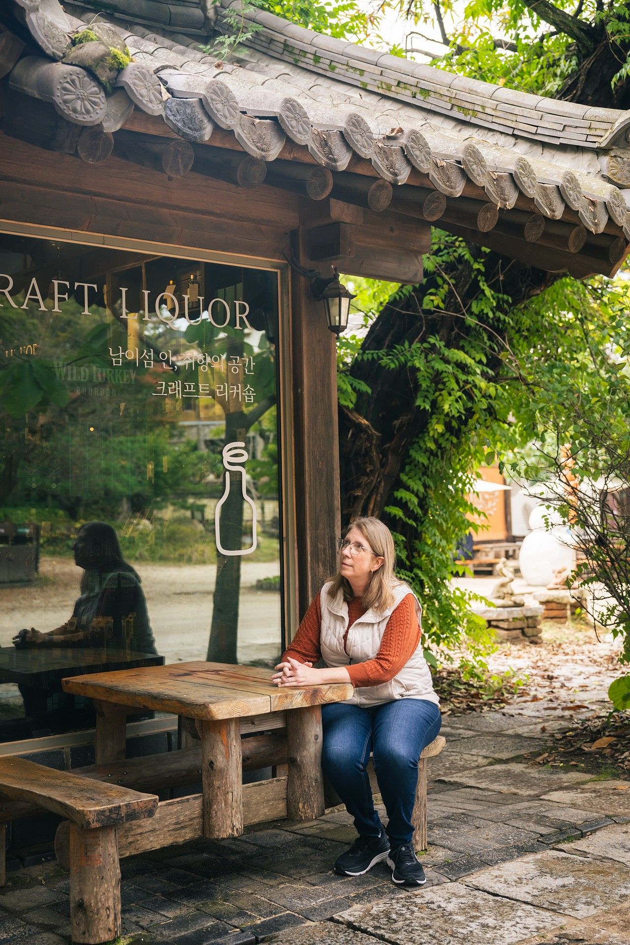 Woman sitting at wooden table outside a Korean craft liquor shop with traditional architecture and lush greenery.
