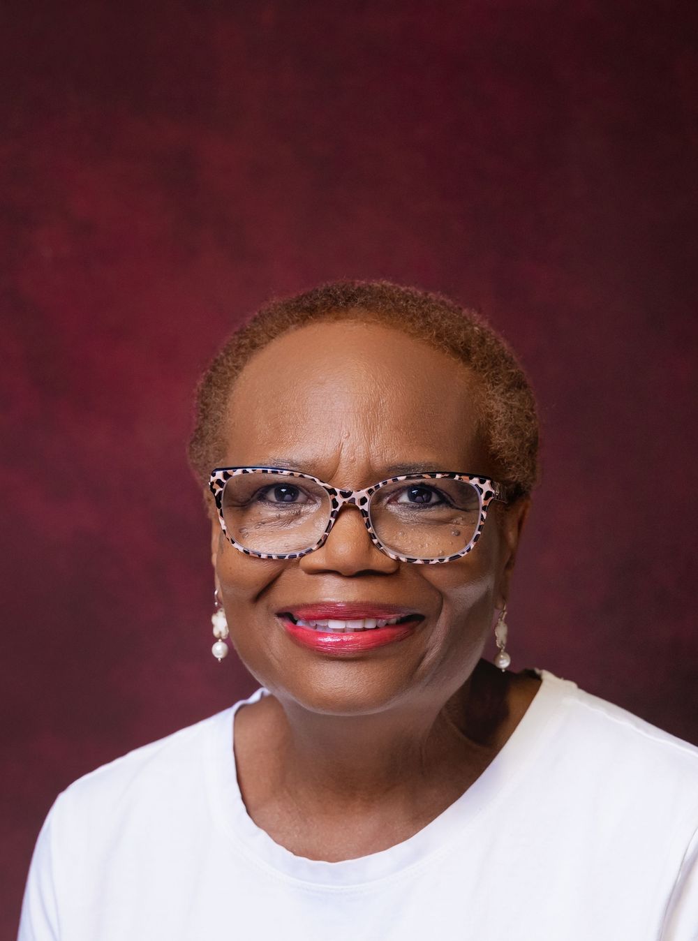 Smiling woman wearing leopard print glasses and white top poses for professional headshot on dark red background.