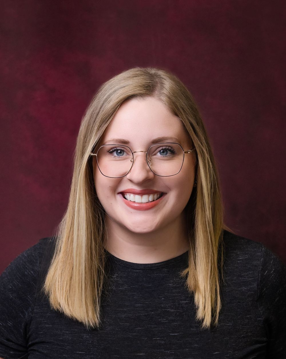 Smiling young woman with blonde hair and rose gold glasses wearing a dark top against a maroon background.