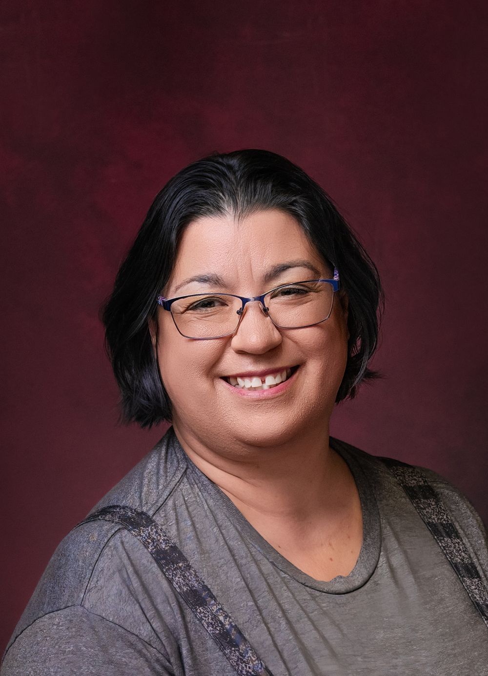 Smiling woman with short dark hair and blue glasses wearing a gray top against a dark red background.