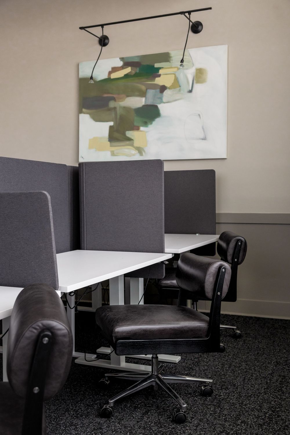 Modern office workspace with black leather chairs, white desks, gray dividers, and abstract green wall art under track lighting.