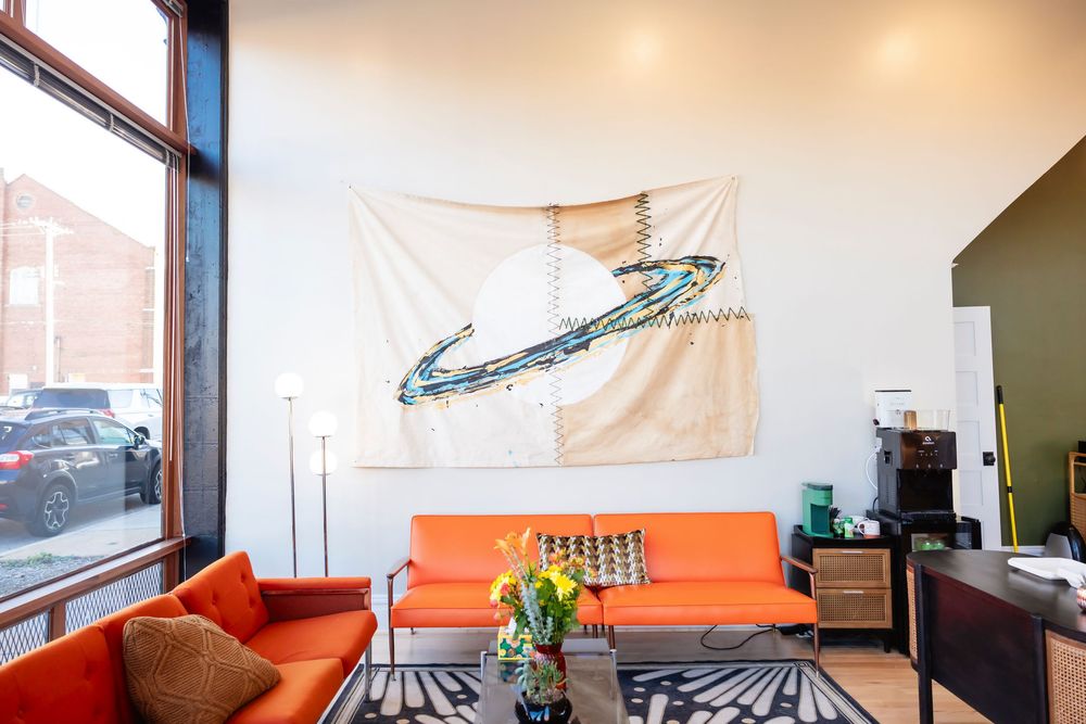 Modern lounge with orange sofas, patterned rug, yellow flowers, and large abstract art banner on white wall.