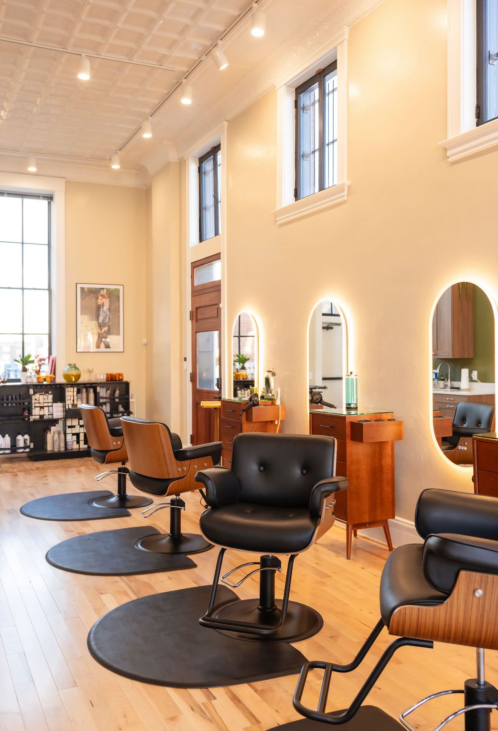 Modern hair salon interior with black styling chairs, arched mirrors, and warm wood accents on light hardwood floors.