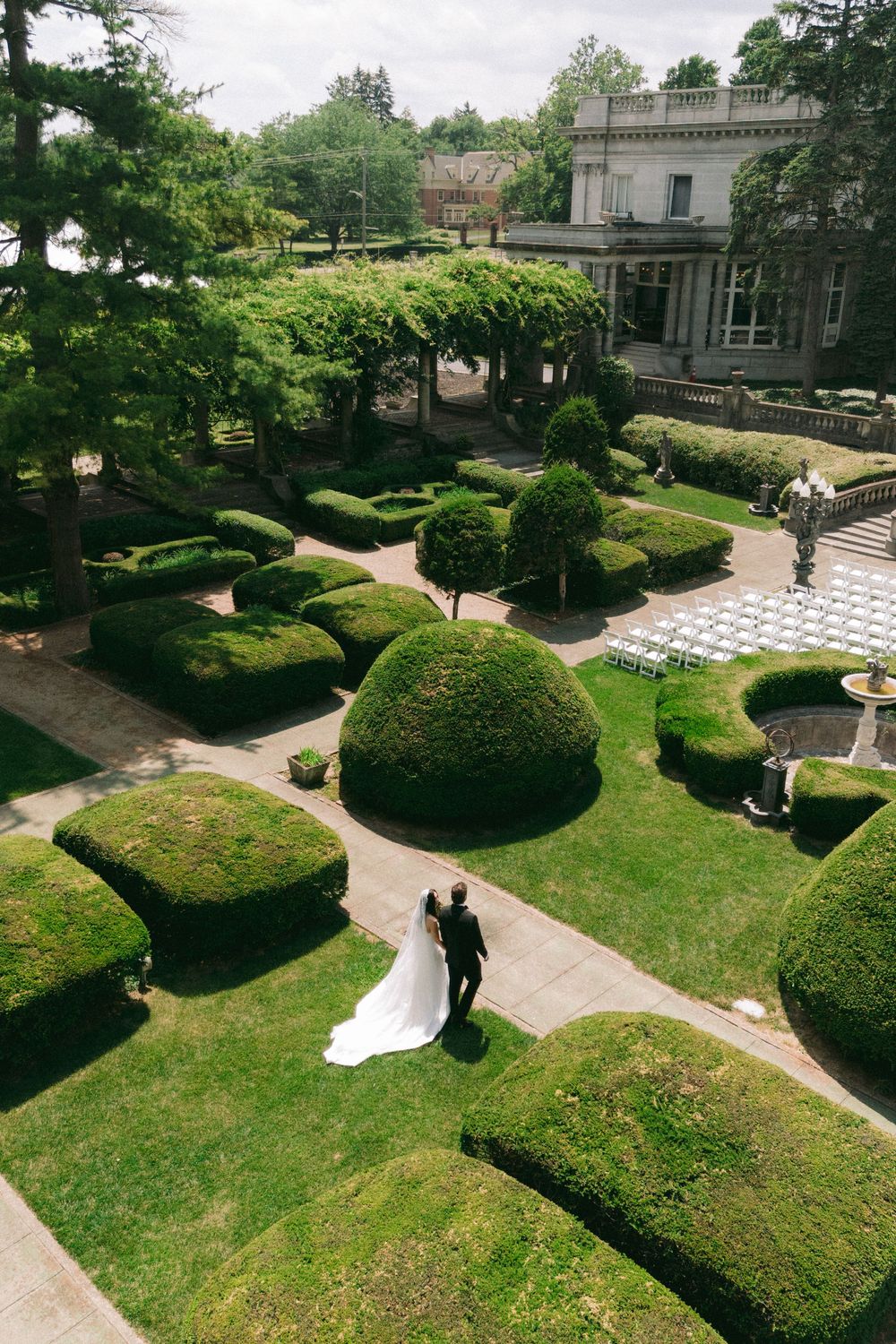 Aerial view of a bride and groom walking through an elegant formal garden with sculpted hedges beside a grand estate.